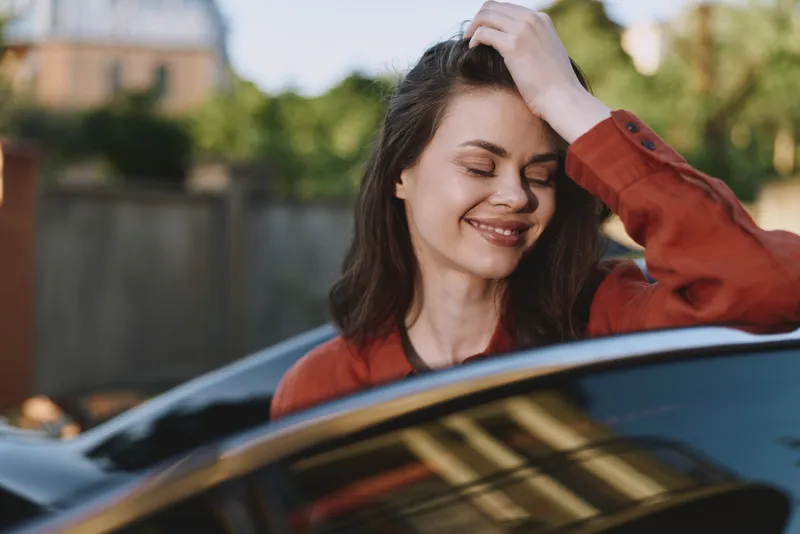 A young woman with shoulder length brunette hair smiles while resting her hand on her head beside a car in a sunny outdoor setting, conveying a carefree lifestyle