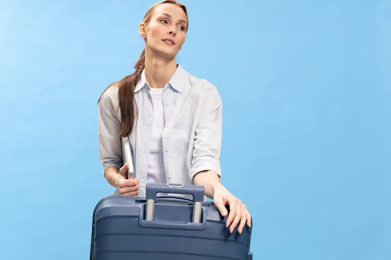 Young woman in casual shirt holding laptop and leaning on suitcase against blue background. Concept for travel, business trip, and modern lifestyle with confident attitude.