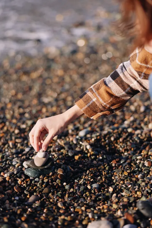 Close up of a hand in plaid sleeve placing a small pebble on a rocky beach, highlighting texture and natural detail with calm water in the background