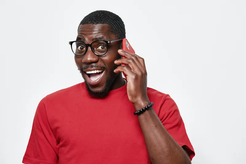 African American man with dark skin wearing glasses and red shirt talking on smartphone with excited expression isolated on white background for communication and technology.