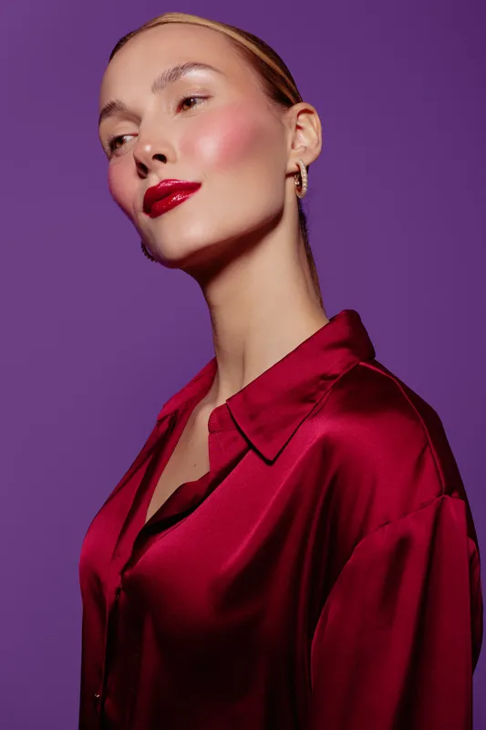 Young woman with glossy red lips and shiny skin wears silky red blouse against purple background. Portrait with elegant makeup and smooth hairstyle. Concept of beauty and fashion.