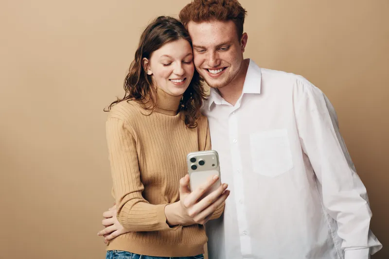 Young couple smiling and taking a selfie together on smartphone. Casual clothing, warm beige background. Concept of friendship, happiness, and modern technology use.