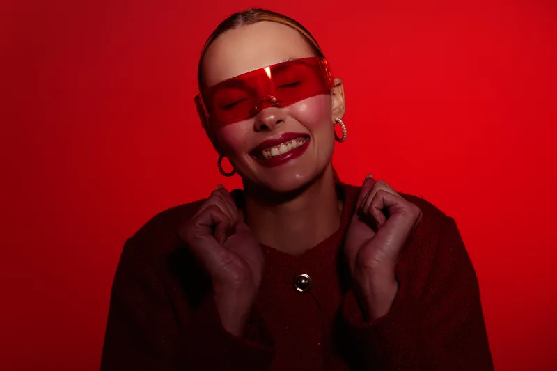 Smiling woman with red transparent glasses and hoop earrings poses happily against a red background. She wears a dark red sweater and expresses joy and confidence in studio lighting.