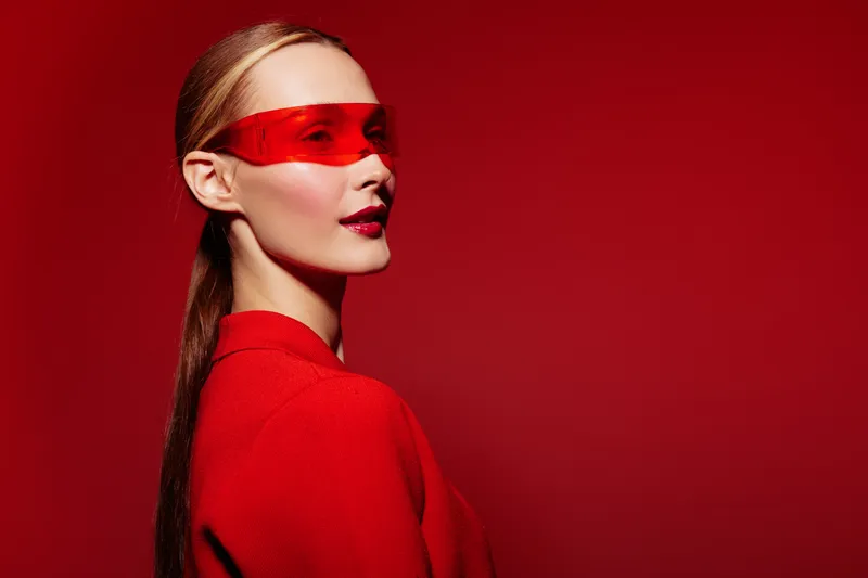 Stylish woman in red outfit and translucent red visor poses against red background. Fashion portrait with sleek hairstyle and bold makeup. Concept of modern style and confident attitude.