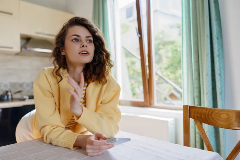 Cheerful woman indoors at a dining table, wearing a casual yellow sweater, natural daylight highlighting a cozy home moment, relaxed posture, candid smile, calm and inviting atmosphere in a bright