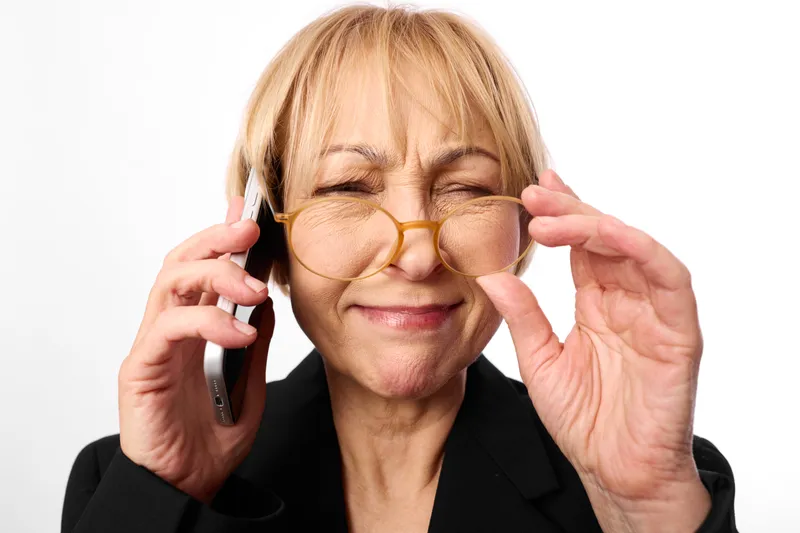 Elderly woman holding a phone to ear, adjusting glasses, eyes strained, mild frustration, vision care concept with daily aging stress on white background