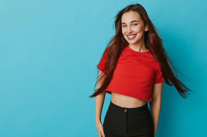 Young woman smiling and posing in red crop top and black pants against blue background. Happy female model with long hair and casual style in studio portrait with confident expression.