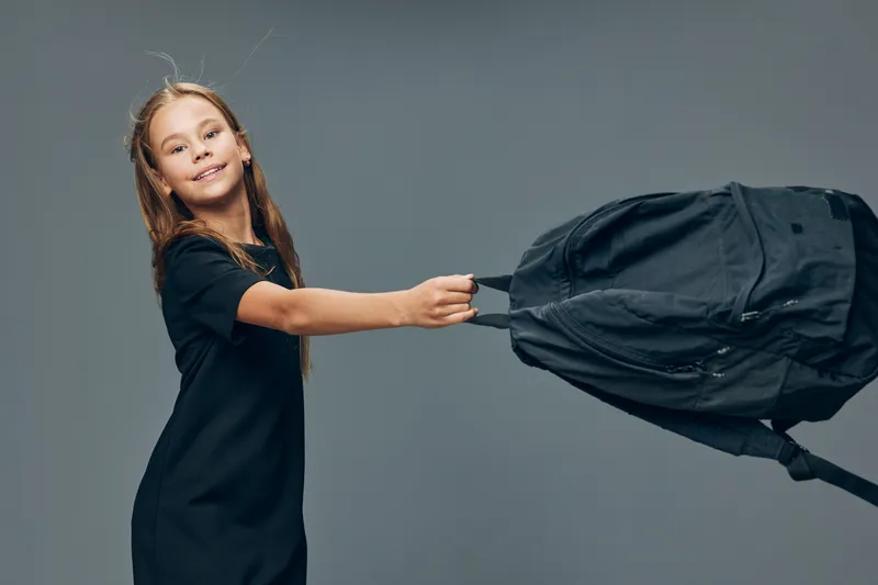Girl in black dress smiling and pulling black backpack strap on gray background with joyful expression and casual posture for youth and school concept.
