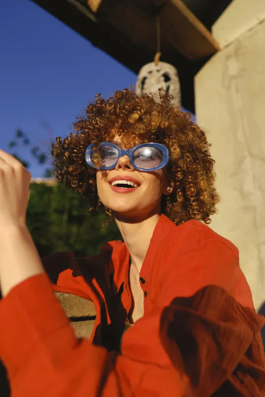 Young woman with curly hair smiles in bright sunlight, wearing a red shirt and oversized blue sunglasses, posing on sunlit steps with a warm glow