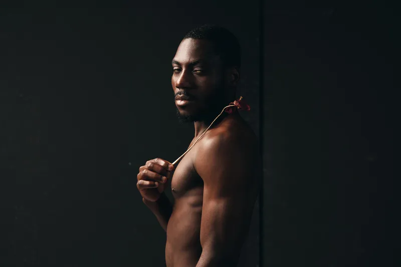 African man with dark skin and muscular body holding a flower on shoulder in creative conceptual visual style. Portrait with dramatic lighting and artistic expression on black background.