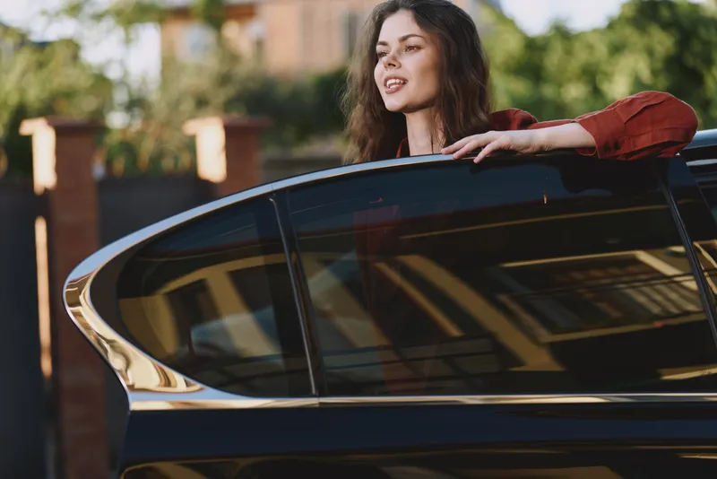 Smiling woman leaning on a car window in an outdoor lifestyle scene, casual fashion and leisure mood with warm daylight and urban backdrop