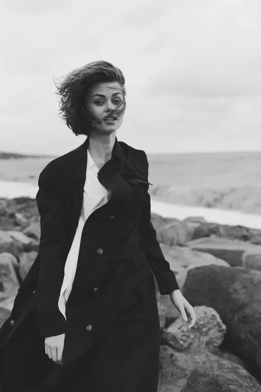 Woman on a windy beach wearing a long coat, dramatic movement and monochrome mood, outdoor fashion beside the sea, wind in hair, serene yet powerful