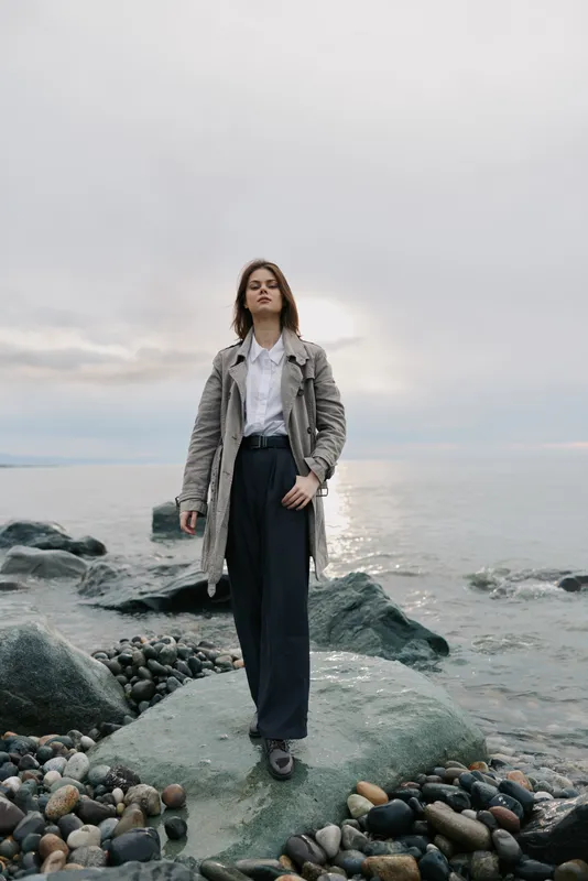 Young woman standing on rocky shore wearing long coat and wide pants. Cloudy sky and calm sea in background. Concept of autumn fashion and outdoor lifestyle.