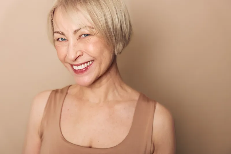 Smiling middle aged woman with short blonde hair wearing beige tank top on neutral background. Concept of happiness, confidence, natural beauty, and mature female lifestyle portrait.