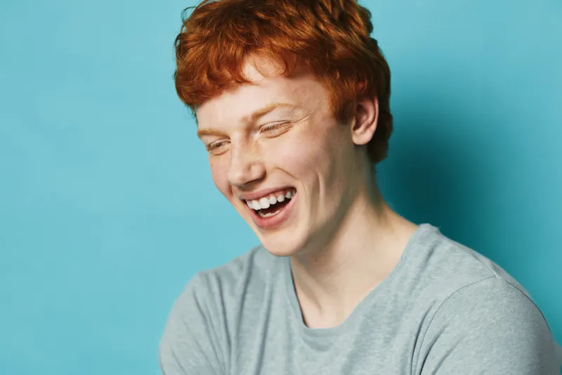 Redhead man laughing with eyes closed against blue background, casual style with gray shirt, happy male portrait and natural smile expression in studio lighting.