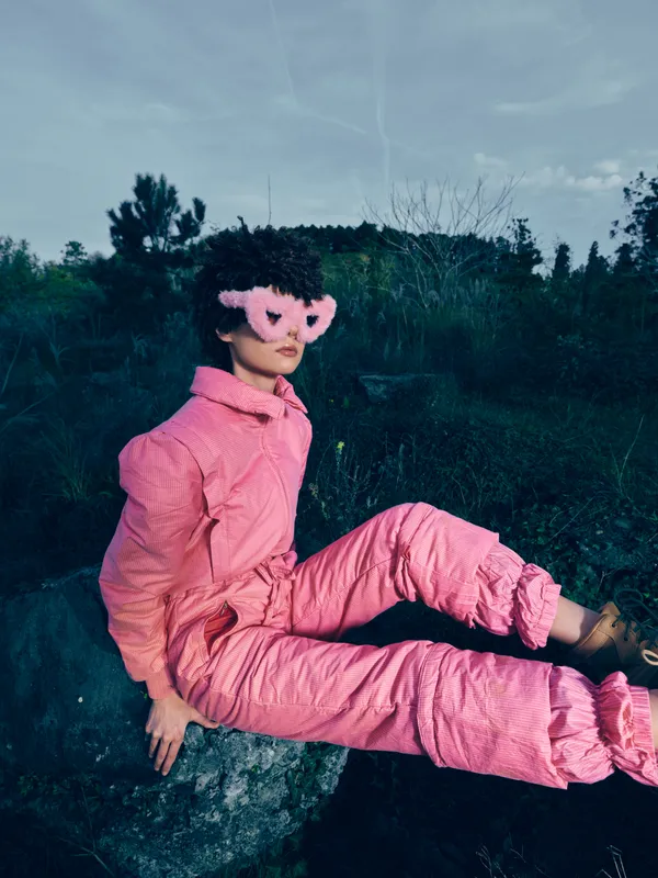 Pink jumpsuit and whimsical mask worn by a person sitting on a rock outdoors, surrounded by trees and blue sky, a bold fashion portrait with a playful mood.