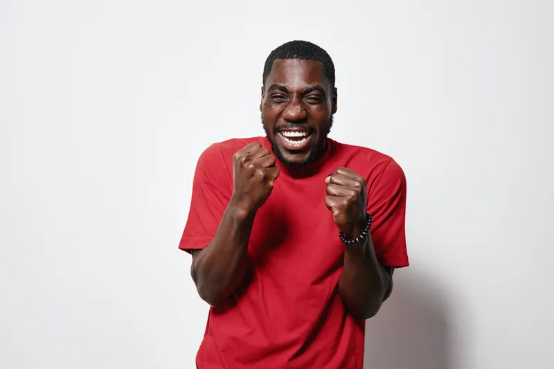 African American man with dark skin wearing red t shirt expressing excitement and joy with clenched fists on white background. Happy black male portrait with positive emotion.