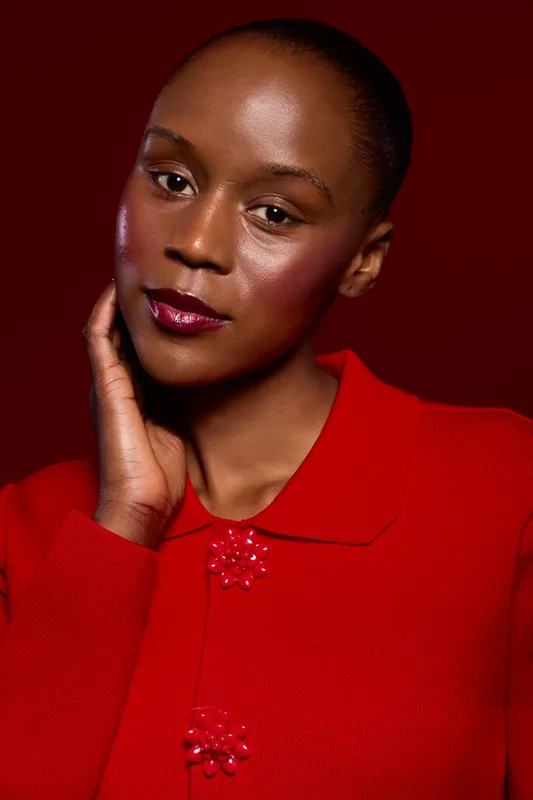 Close-up portrait of a confident black woman with dark skin wearing a red sweater against a dark red background. The concept highlights beauty, fashion, and elegance with natural makeup.