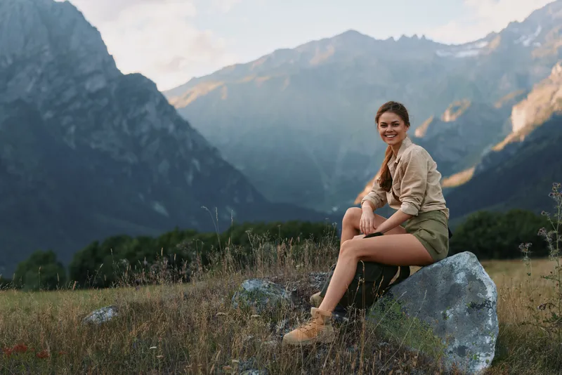 Young woman with long hair sits on a rock in a mountain meadow smiling and enjoying the natural landscape during a calm late afternoon.