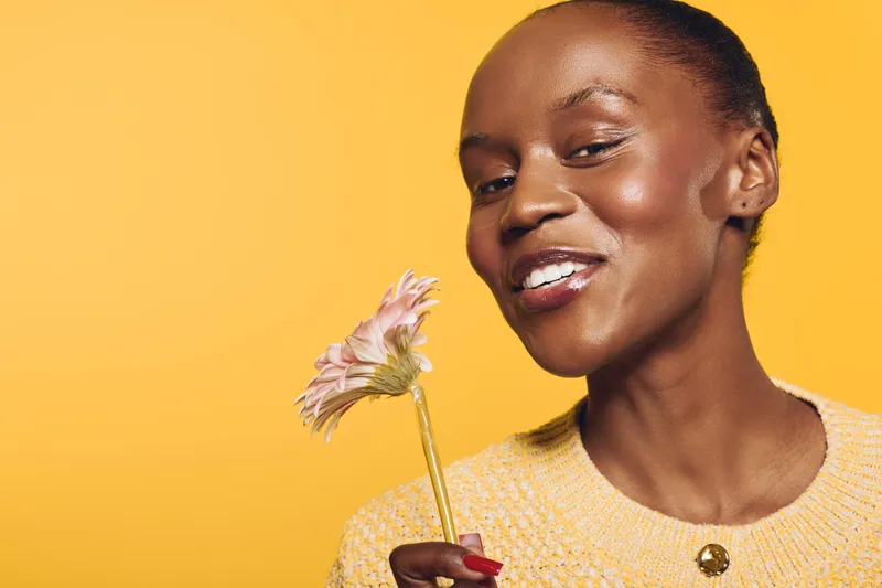 Smiling African American woman with dark skin holding pink flower on yellow background. Concept of beauty, happiness, natural skincare, and positive mood in studio portrait photography.