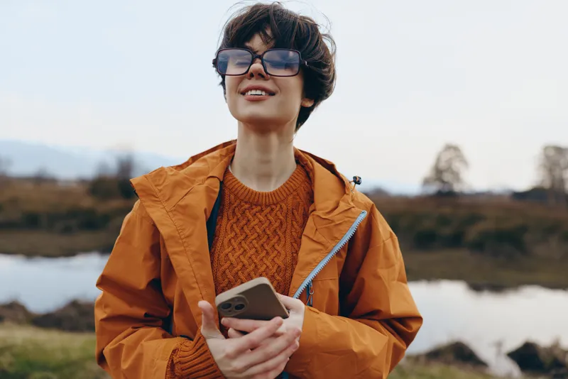 Woman lifestyle outdoors, orange jacket and sunglasses, smiling while holding smartphone near a lake, natural scenery, fresh air, casual travel, daylight, blurred background, open hills view