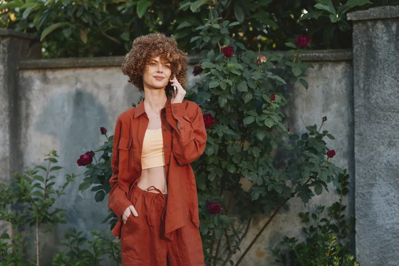 Woman lifestyle portrait outdoors in a rust jacket and beige top, standing among roses with a confident, casual streetwear vibe and natural light, relaxed candid mood, sunny