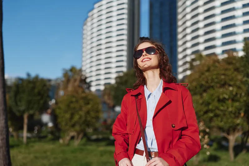 Young woman in a red coat and sunglasses enjoys sunny weather while walking in an urban park with modern buildings in the background.