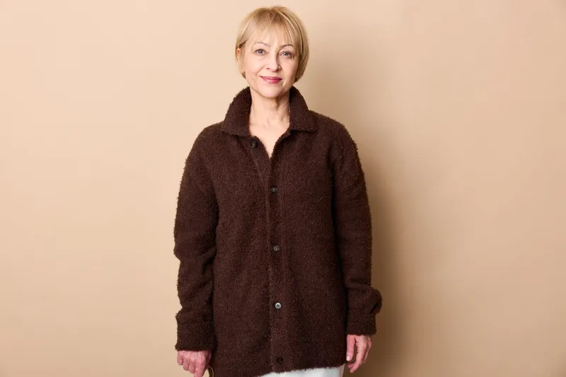 Elderly woman standing in a studio, beige background, wearing dark wool coat, calm portrait with hands relaxed, indoor lifestyle concept, soft light.