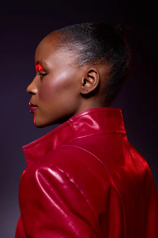 Profile of a dark skinned African woman with glossy skin and bright red makeup wearing a shiny red leather jacket. Studio portrait with dramatic lighting and modern fashion concept.