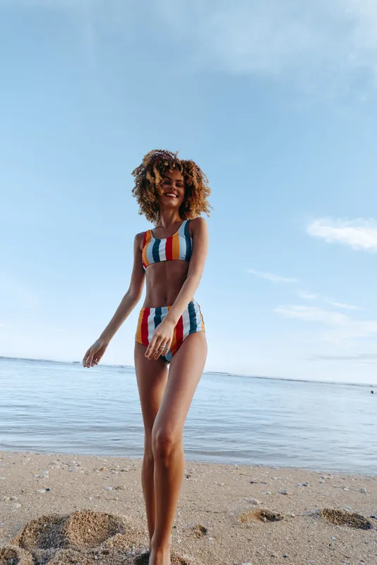Young woman with curly hair walks along the sandy beach in a colorful striped swimsuit under a clear blue sky, enjoying a sunny summer day by the calm sea.