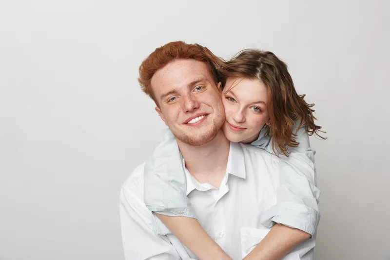 Smiling young couple with red hair and casual clothes embraces on white background. Happy man and woman show affection and closeness in studio portrait. Concept of love and friendship.