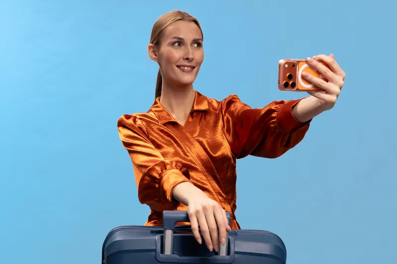 Young woman in orange blouse takes selfie with smartphone while holding suitcase handle. Blue background, travel concept for vacation planning and trip preparation.