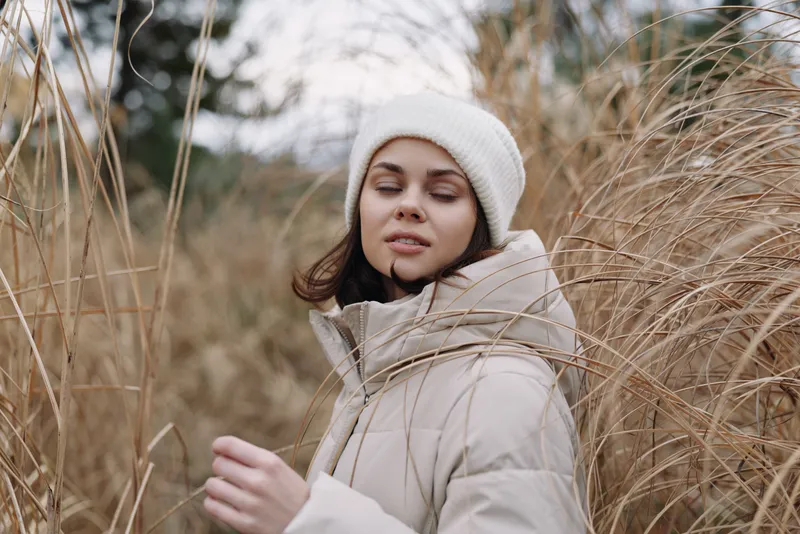 Woman in a winter coat and beanie stands among tall grasses in an open field, serene expression and soft light creating a calm outdoor portrait, natural mood.