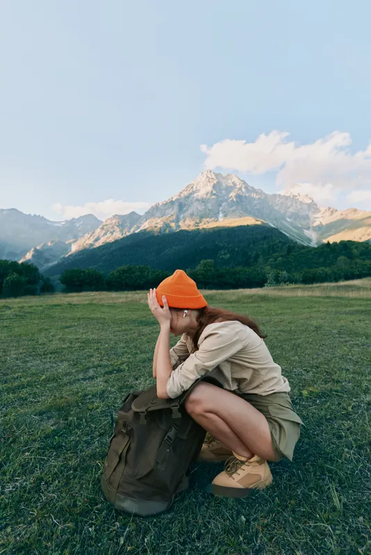 Young woman crouches in a sunlit meadow with a backpack, shielding her eyes as rugged mountains rise in the background.
