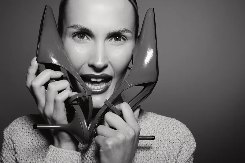 White woman with fair skin holds high heel shoes near face, smiling playfully. Studio portrait in black and white, concept for fashion, footwear, and joyful expression.