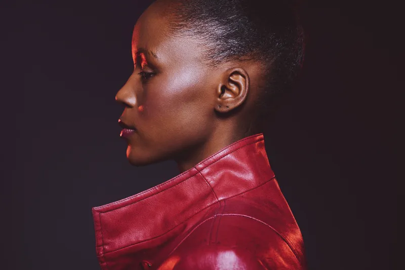 Profile of an African woman with dark skin wearing a red leather jacket against a dark background. Concept of fashion, beauty, confidence, and elegance in studio portrait.