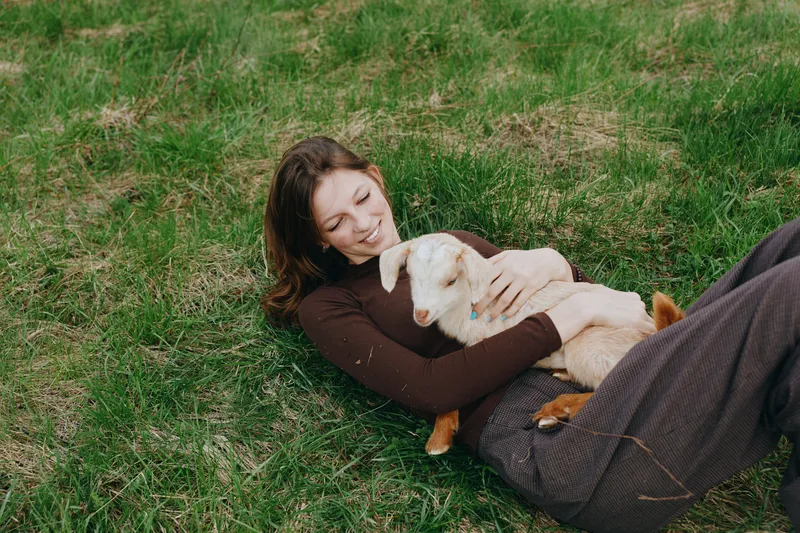 Cheerful woman outdoors, lying on grass while embracing a gentle goat kid, warm sunlight, relaxed pose, candid moment, pastoral setting, soft breeze, natural tones, gentle affection
