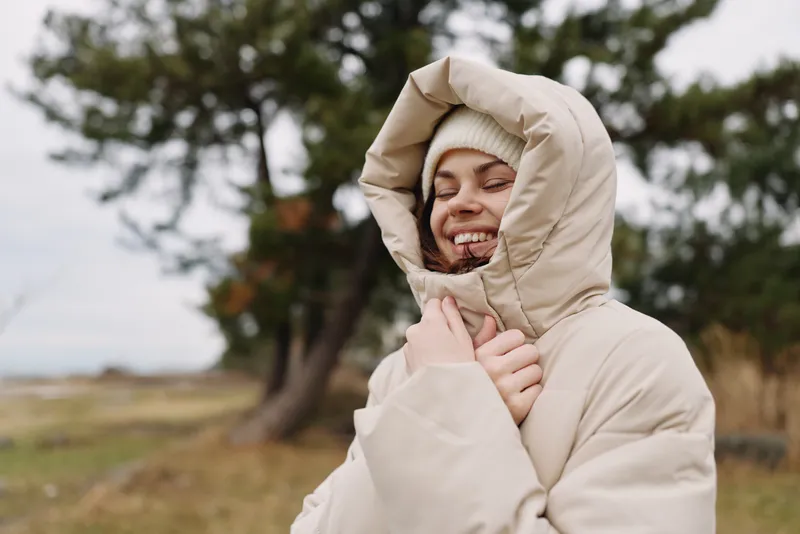 Woman in a puffy hooded coat outdoors, smiling warmly amid trees and an open field, in chilly weather with a cozy wind.