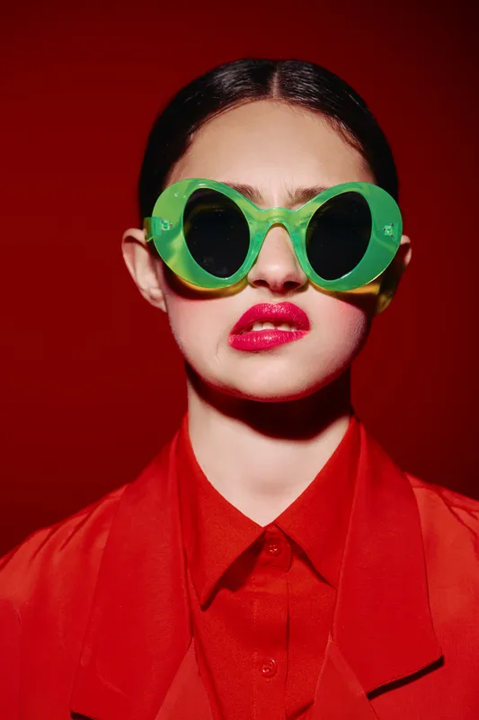 Fashion model wearing oversized green round sunglasses and vivid red lipstick with a glossy finish. Bright red blazer contrasts with vibrant background. Bold beauty creative concept.