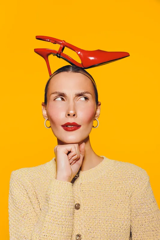 Thoughtful woman with red lipstick and hoop earrings balances red high heel shoe on head against bright yellow background. Concept for fashion, creativity, and style expression.
