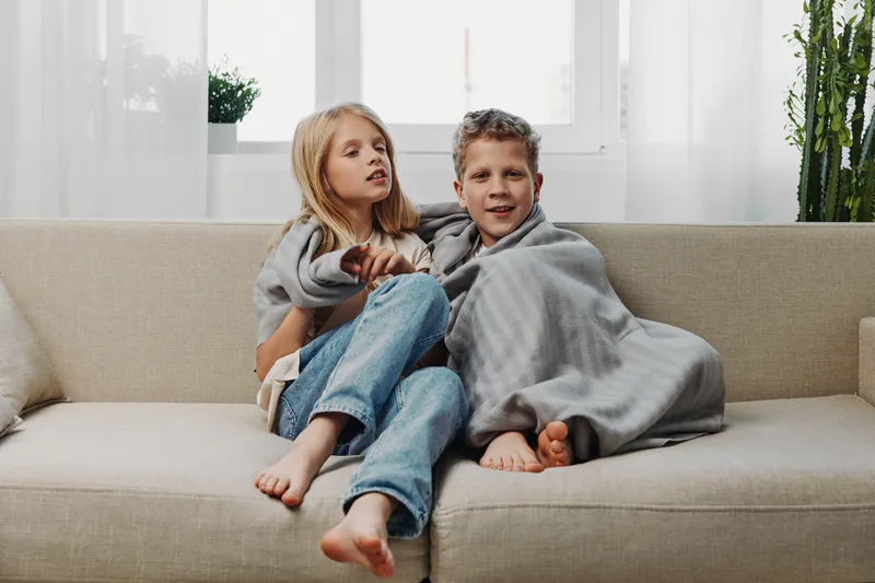Two children wrapped in gray blanket sitting barefoot on beige sofa by bright window in cozy living room. Concept of warmth, comfort, and sibling bonding.