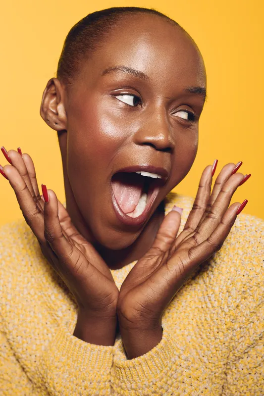 Happy young black woman with dark skin wearing yellow sweater poses with open mouth and hands near face on yellow background. Concept of joy, excitement, and emotional expression.