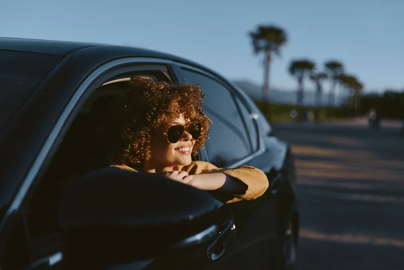 Young woman with curly hair leans out of a car window, smiling as palm trees glow in warm retro sunlight, hinting at carefree road trip vibes and nostalgic film tones.