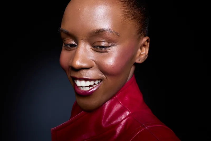 Close-up of happy African woman with dark skin wearing red leather jacket smiling with eyes closed against black background. Concept of joy, fashion, beauty, and confident female model.