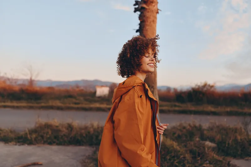 Woman lifestyle outdoors, curly hair, tan jacket, stands in a countryside field at sunset, looking ahead in soft evening light, calm natural portrait with road and open sky background