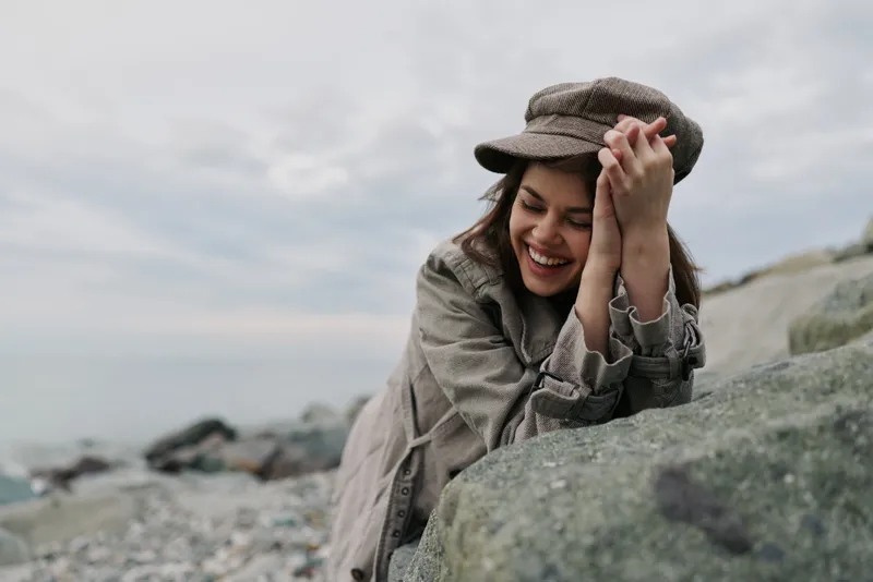 Smiling woman in vintage outfit leans on rock near rocky beach. Overcast sky creates moody atmosphere. Concept for happiness, nature, fashion, and outdoor lifestyle.