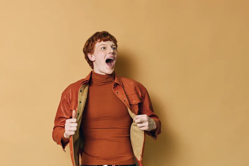 Young man with red hair wearing orange jacket and turtleneck looking excited with open mouth against plain beige background. Expression of surprise and enthusiasm.