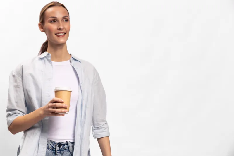Smiling woman in casual clothes holding a takeaway coffee cup looks to the side with a relaxed expression. Bright studio background creates a fresh and positive mood.