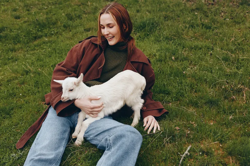 Smiling woman sits on grass outdoors, cradling a young goat in her arms. Warm, relaxed lifestyle scene with natural light and rustic countryside mood. A warm, affectionate moment now