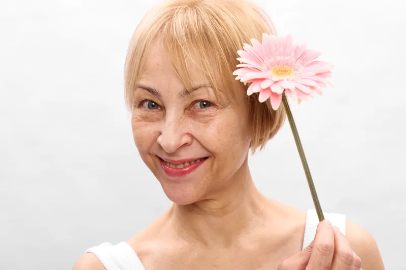 Woman portrait, smile, elderly blonde holding pink flower on white studio background, gentle expression, soft light, minimal setting, close up headshot, calm mood, beauty, tenderness
